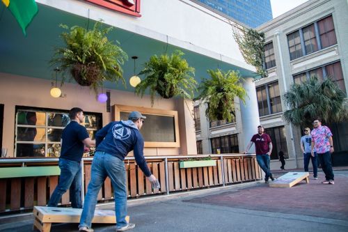 Two people playing cornhole outside a building, with greenery and urban scenery in the background.