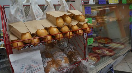 A display of fresh bread rolls in paper bags, with additional loaves in plastic wrap, next to a refrigerated case.