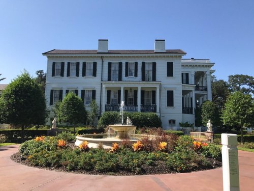 A large white mansion with black shutters, surrounded by lush greenery and a fountain in a landscaped garden.