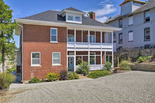 A two-story brick house with a porch, surrounded by greenery and gravel, set against a clear blue sky.