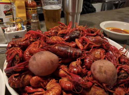 A large platter of boiled crawfish with two potatoes, accompanied by a beer and condiments in the background.