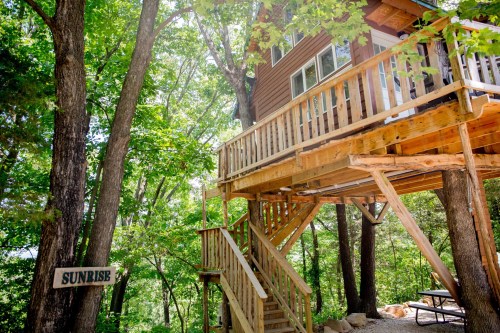 A wooden treehouse with a balcony, surrounded by lush green trees, and a sign reading "Sunrise" nearby.