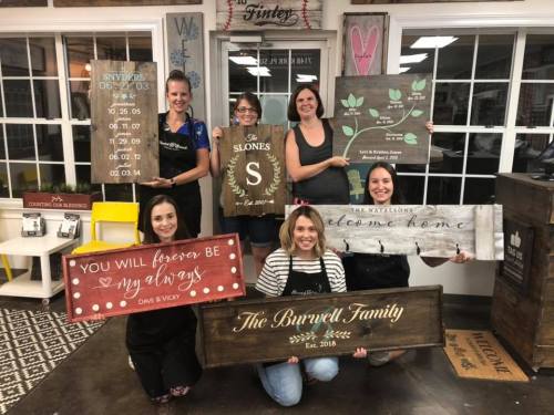 A group of six women holding personalized wooden signs in a craft studio, smiling and posing together.