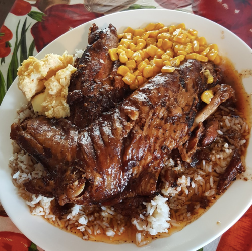 A plate of rice topped with seasoned meat, corn, and mashed potatoes, served on a colorful floral tablecloth.