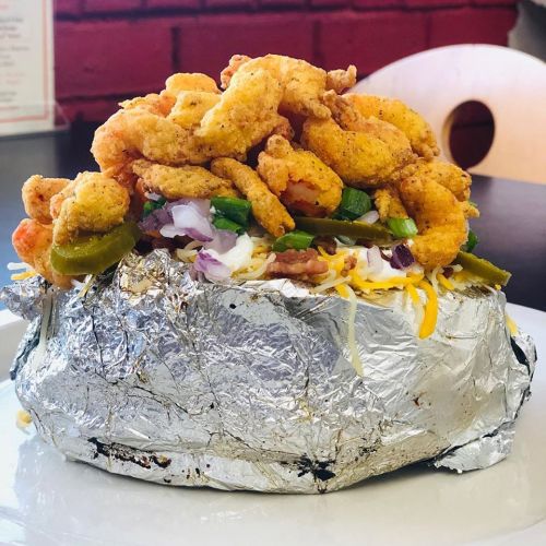 A large burrito wrapped in foil, topped with crispy shrimp, jalapeños, onions, and cheese on a white plate.