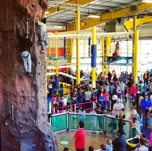 A busy indoor climbing gym with people climbing a rock wall and others watching, surrounded by colorful structures.