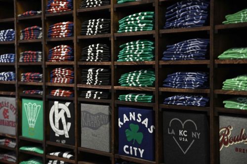 A display of neatly folded t-shirts in various colors, featuring different logos and designs on wooden shelves.