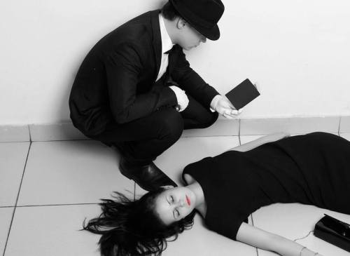 A man in a suit and hat kneels beside a woman in a black dress, who lies on the ground with red lipstick.