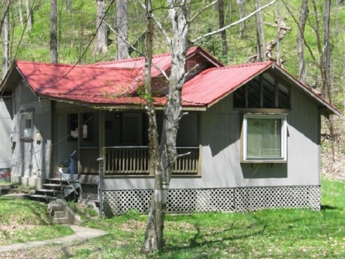 A small, rustic house with a red roof, surrounded by greenery and trees in a wooded area.