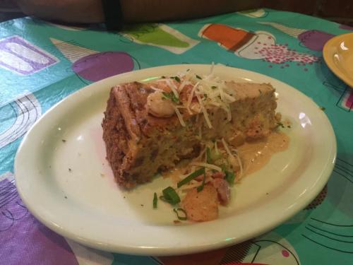 A slice of savory seafood cake topped with shrimp and cheese, served on a white plate against a colorful tablecloth.
