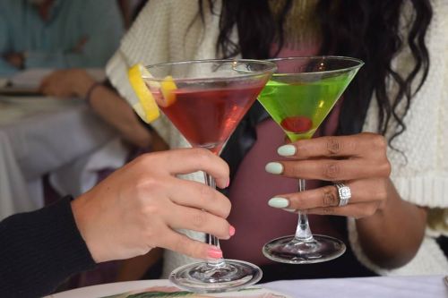 Two hands clink cocktail glasses, one with a red drink and lemon slice, the other with a green drink and cherry.