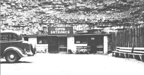 Black and white photo of a cave entrance with a sign, an old car parked nearby, and wooden benches in front.