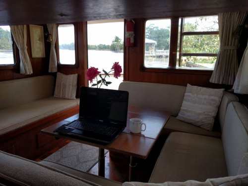 Cozy interior of a boat with a laptop on a table, a coffee mug, and flowers by the window overlooking the water.