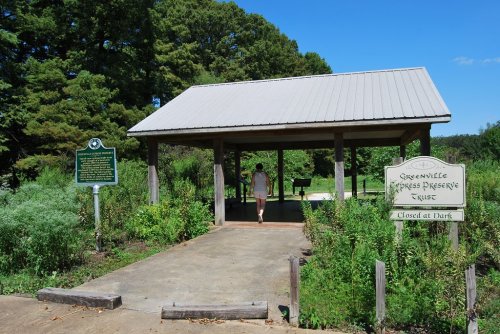 A person walks towards a covered pavilion in a park, surrounded by greenery and informational signs.