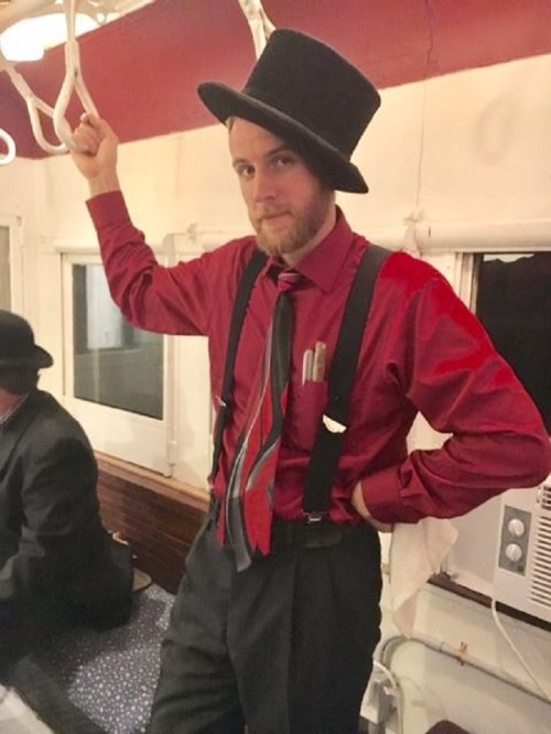 A man in a top hat and red shirt stands confidently, holding onto a rail in a vintage train setting.