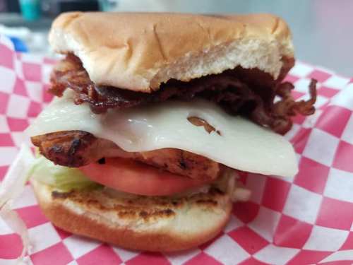 A close-up of a sandwich with grilled chicken, melted cheese, bacon, lettuce, and tomato on a toasted bun.