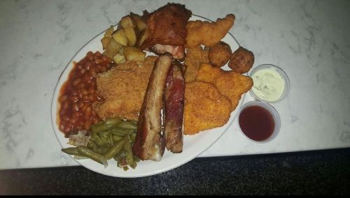 A large plate of assorted fried foods, including chicken, fish, ribs, green beans, baked beans, and potato wedges.