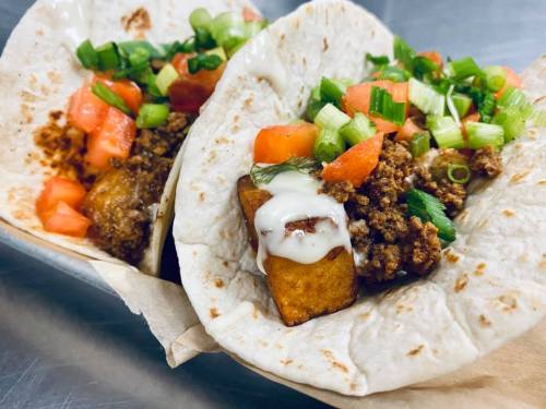 Two soft tortillas filled with seasoned meat, diced tomatoes, green onions, and a drizzle of sauce.