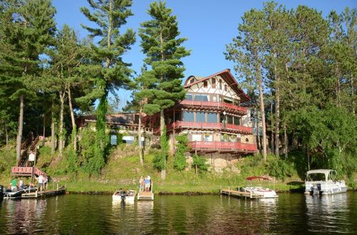 A large, rustic house surrounded by trees, overlooking a calm lake with boats docked nearby.