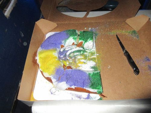 A partially sliced king cake with purple, green, and yellow sprinkles in a cardboard box, next to a knife.