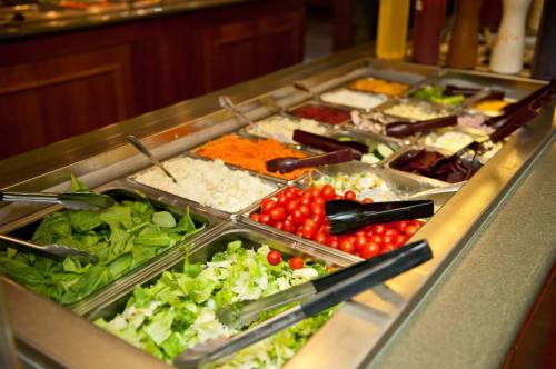 A salad bar featuring various toppings, including greens, tomatoes, cheese, and dressings, with serving utensils.