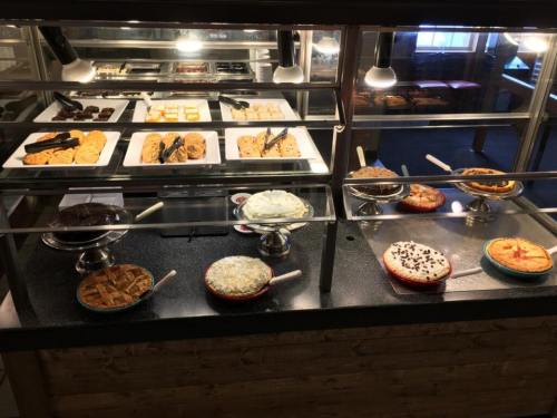 A dessert display featuring various cakes, pies, and cookies on glass shelves with serving utensils.