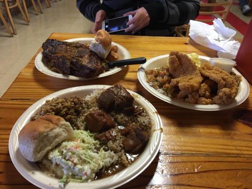 A wooden table with three plates of food: grilled meat, fried fish, rice, coleslaw, and a bun. A person is using a phone.