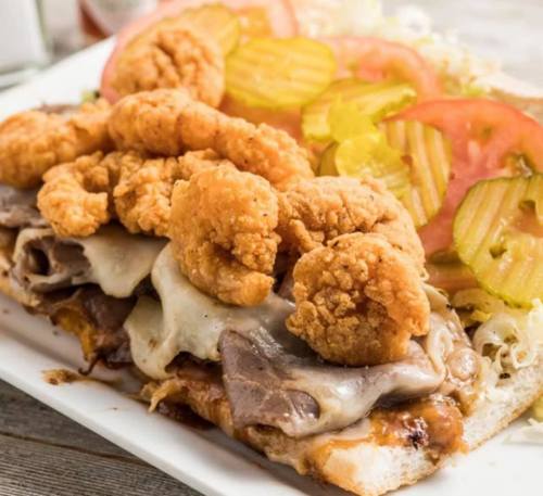 A hearty sandwich with roast beef, melted cheese, fried shrimp, lettuce, tomatoes, and pickles on a toasted bun.