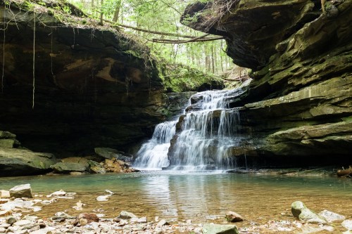 A serene waterfall cascades over rocks into a clear pool, surrounded by lush greenery and steep cliffs.