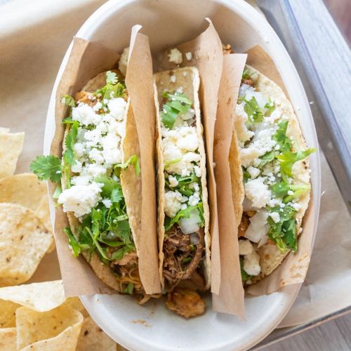 Three tacos filled with meat, onions, and cilantro, served in a bowl with tortilla chips on the side.