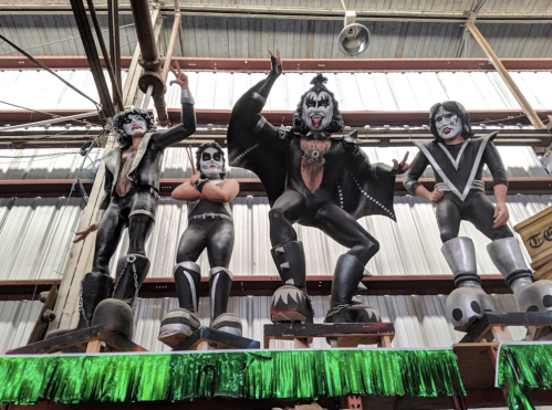 Four large, colorful figures resembling rock band members, dressed in black and silver costumes, on display in a warehouse.