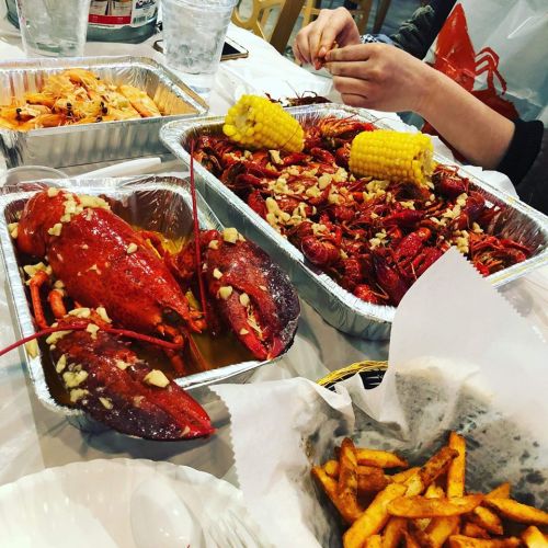 A table with seafood dishes, including lobsters, crawfish, corn, and fries, surrounded by drinks and utensils.