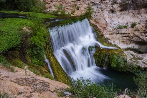 A serene waterfall cascades over rocks, surrounded by lush greenery and a tranquil pool below.