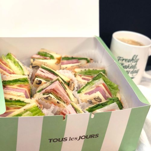 A box of colorful sandwiches with layers of ham, lettuce, and cheese, next to a coffee mug.
