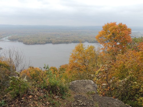 A scenic view of a river surrounded by autumn foliage, with vibrant orange and yellow trees under a cloudy sky.