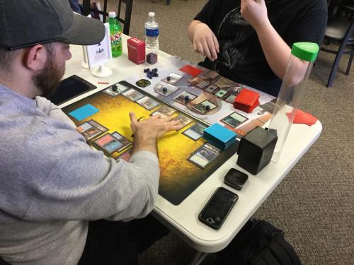 Two players engaged in a card game at a table, with various game pieces and drinks nearby.