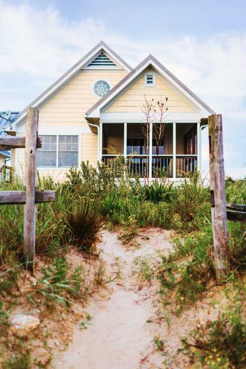 A yellow beach house surrounded by greenery, with a sandy path leading to its porch under a blue sky.