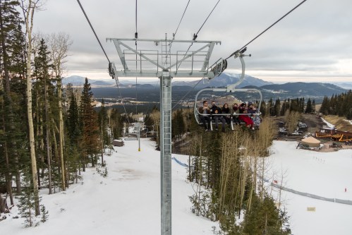 A group of six people on a ski lift, surrounded by snow-covered trees and mountains in the background.