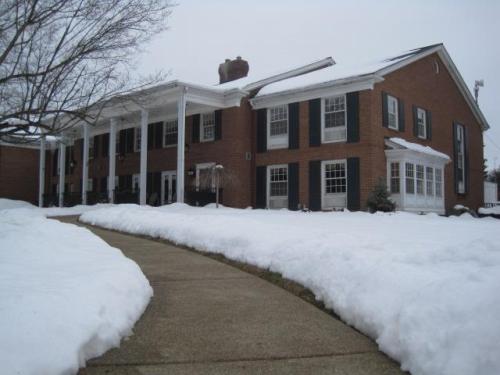 A brick building with white columns and green shutters, surrounded by snow and a curved walkway on a cloudy day.