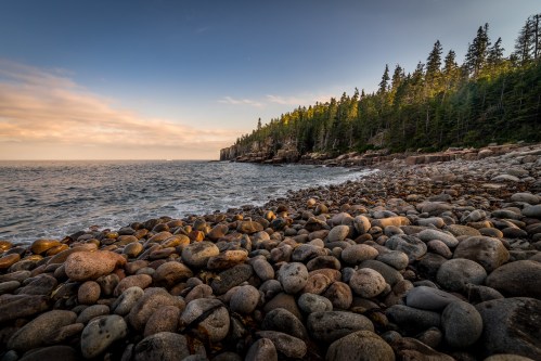 A rocky shoreline with smooth pebbles, surrounded by trees and a calm ocean under a clear sky.