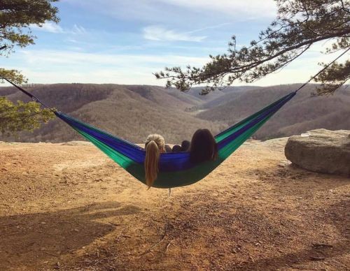 Two people relax in a hammock overlooking a scenic valley with rolling hills and trees under a clear sky.