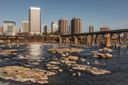 A city skyline with modern buildings overlooks a river with rocky islands and a bridge in the foreground.