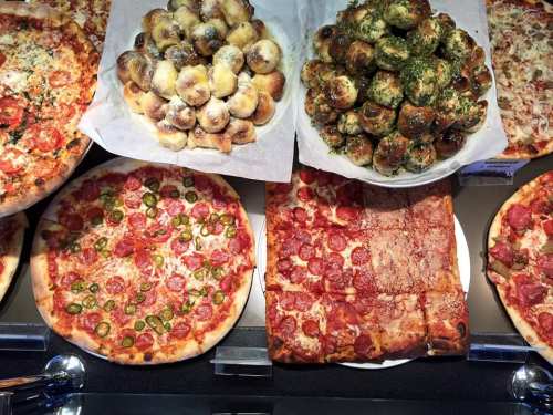 A display of various pizzas, including pepperoni and jalapeño, alongside garlic knots and cheesy bread bites.