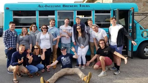 A group of people poses joyfully in front of a blue bus for an all-inclusive brewery tour.