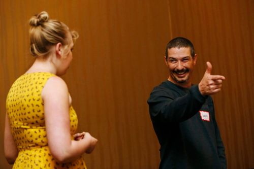 A man smiles and gestures while talking to a woman in a yellow dress at an event.