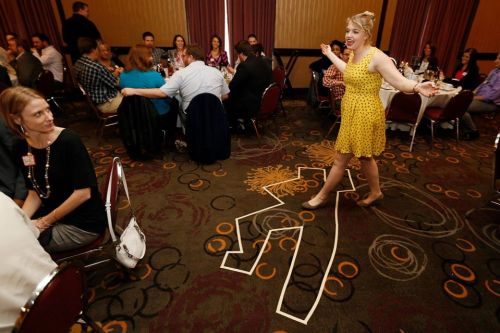 A woman in a yellow dress performs near a chalk outline on the floor, surrounded by a dining crowd.