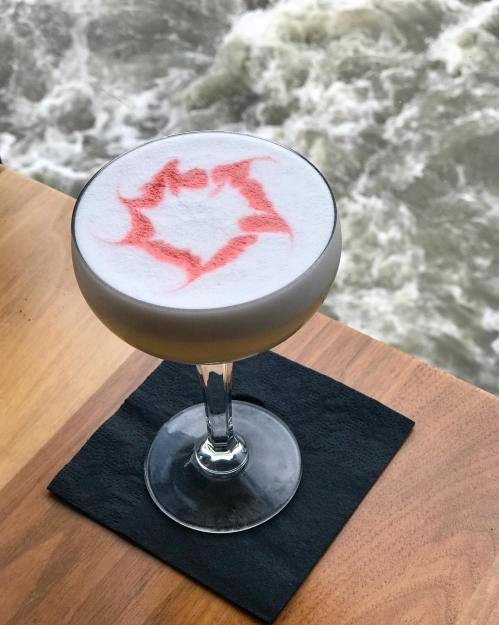 A cocktail in a coupe glass with a frothy top and red design, set on a wooden table near flowing water.