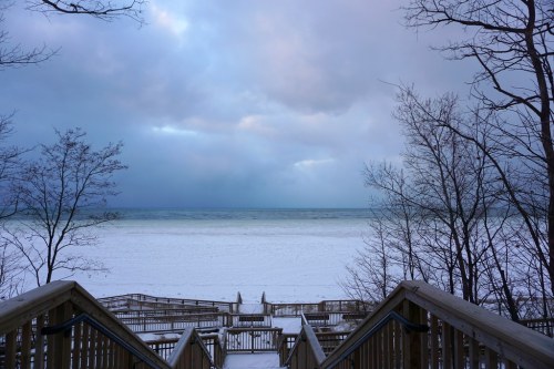 A snowy beach scene with wooden stairs leading down to the shoreline, under a cloudy sky.
