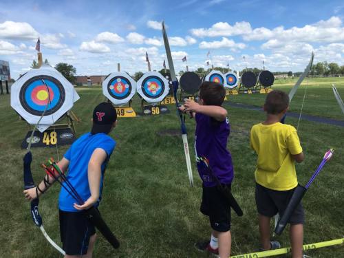 Three children aiming bows at colorful archery targets on a sunny day, with blue skies and grass in the background.