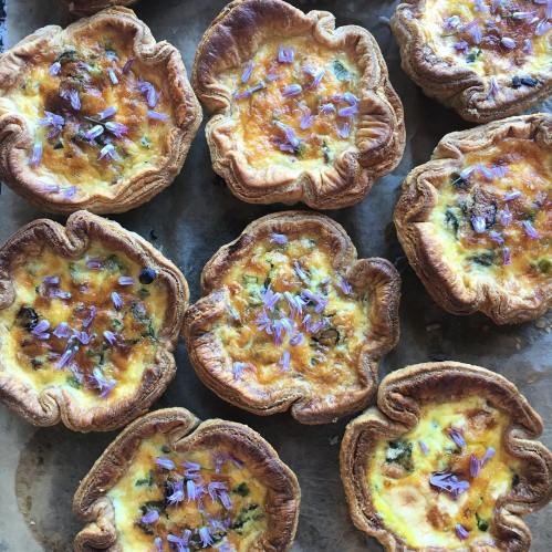 A tray of golden-brown mini quiches topped with purple flowers, arranged neatly on parchment paper.